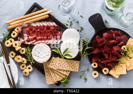 Salumi e formaggi sono serviti su un vassoio su un tavolo con il vino bianco, crackers, grissini e taralli con erbe aromatiche su un lino blu tavola festiva Foto Stock