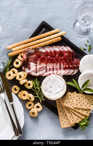 Salumi e formaggi sono serviti su un vassoio su un tavolo con il vino bianco, crackers, grissini e taralli con erbe aromatiche su un lino blu tavola festiva Foto Stock