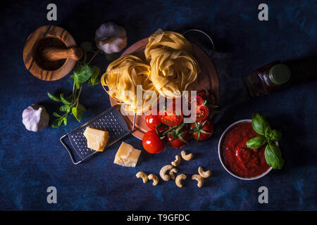 La tabella con gli ingredienti per fare il pesto di pomodoro. Pomodori, aglio, origano fresco e basilico erbe, bottiglia di olio d'oliva, alcune noci di acagiù, parmigiano, c Foto Stock