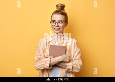 Felice giovane bella donna con hairbun permanente e la holding tablet su sfondo giallo e cercando in disparte . istruzione il concetto di spazio di copia Foto Stock