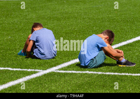 Istruzione, bullismo, conflitto, le relazioni sociali e il concetto di persone - due triste deluso ragazzi seduta di schiena sull'erba nel campo dello stadio Foto Stock