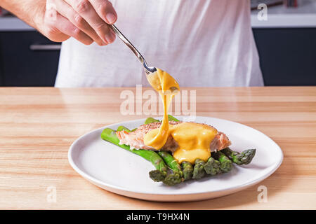 L'uomo le mani versando salsa olandese sulla sommità del delizioso salmone cotto al vapore con asparagi verdi. Presentazione di alimentare il processo di preparazione Foto Stock