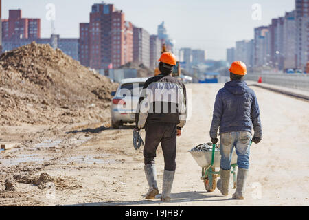 Due lavoratori nei caschi guidare una carriola contro lo sfondo di edifici di nuova costruzione. Foto Stock