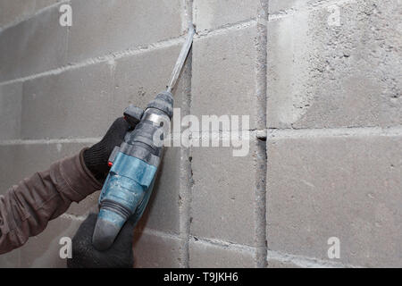 Le mani del lavoratore in guanti tagliati attraverso la parete in calcestruzzo con un perforatore, close-up. Foto Stock