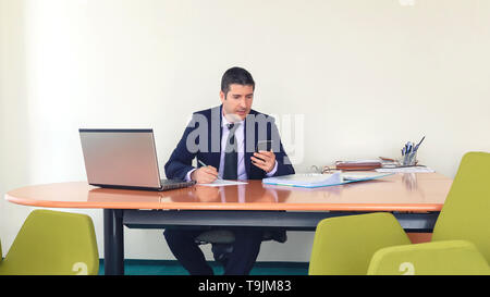 Fiducioso business man digitando sullo smartphone e la scrittura di un documento mentre si è seduti alla scrivania in ufficio - imprenditore maturo in abiti formali usando il telefono Foto Stock