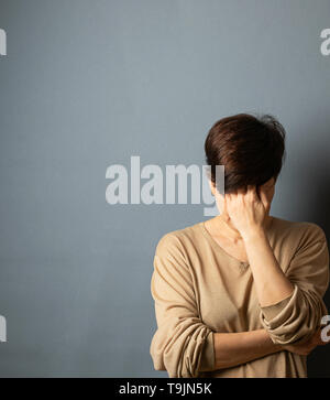 Una donna si copre la faccia con la sua mano, si erge contro il muro. Ritratto di donna su sfondo grigio. Foto Stock