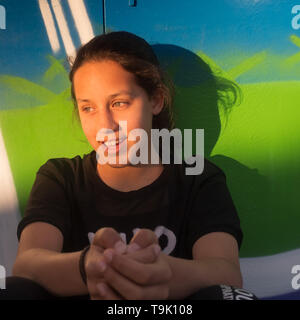 Gioiosa teen ragazza è in appoggio, appoggiata contro un muro di ferro verniciata con colori intensi. Ella è rilassante nella strada all'aperto al tramonto, seduto da solo e Foto Stock