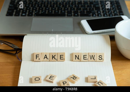 Parola chiave concettuale fake news in piastrelle in legno lettere in casa personale impostazione scrivania con computer portatile, notebook e accessori Foto Stock