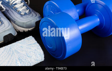 Un paio di scarpe da ginnastica, due manubri blu e un asciugamano al buio su un sfondo di legno Foto Stock