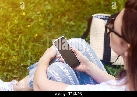 Donna che mantiene il telefono cellulare con schermo scuro nella sua mano. Sfondo di erba, raggi solari. Messa a fuoco selettiva. Foto Stock