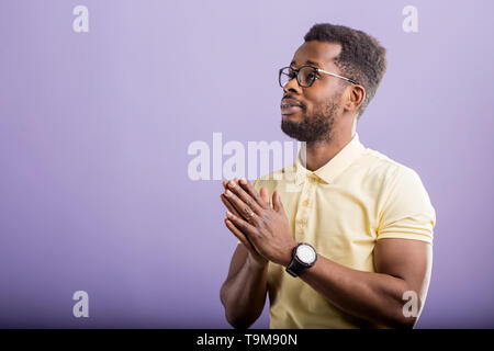 Closeup Ritratto di giovane americano africano uomo con piccola barba, indossando occhiali, polo gialla di t-shirt, guardando a lato, tenendo le palme insieme, prayin Foto Stock