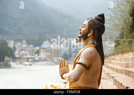 Splendida vista di una seduta statua indù sulla sponda del fiume Gange. Sfocato Lakshman Jhula ponte in background. Foto Stock