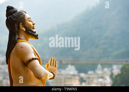 Splendida vista di una seduta statua indù sulla sponda del fiume Gange. Sfocato Lakshman Jhula ponte in background. Foto Stock