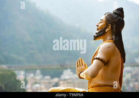 Splendida vista di una seduta statua indù sulla sponda del fiume Gange. Sfocato Lakshman Jhula ponte in background. Foto Stock