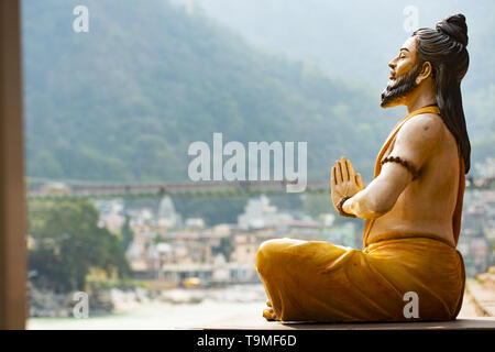 Splendida vista di una seduta statua indù sulla sponda del fiume Gange. Sfocato Lakshman Jhula ponte in background. Foto Stock