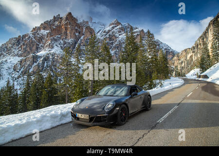 La costosa auto sportiva si ferma sulla strada torsata delle Dolomiti, monumento dell'UNESCO, montagne innevate e alberi verdi sullo sfondo Foto Stock