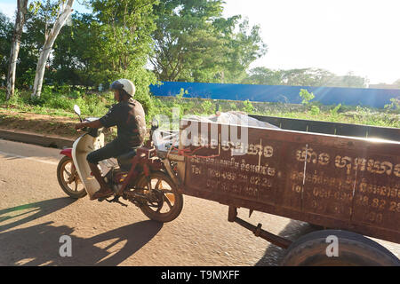 Uomo alla guida di moto durante il traino di un rimorchio di grandi dimensioni in un assolato pomeriggio estivo vicino a Siem Reap, Cambogia. Foto Stock