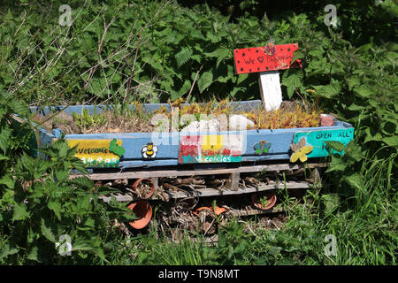 Bug hotel composto da materiali riciclati pallet di legno da una siepe in un ambito comunitario in primavera con erba, ortiche e di altri tipi di vegetazione crescente intorno ad esso. Foto Stock