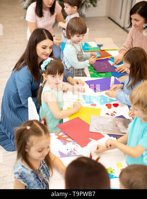 Prechool childs lavora con carta sotto la supervisione degli insegnanti. Un gruppo di bambini piccoli facendo del progetto in una scuola materna. Kids artigianale in aula. Foto Stock