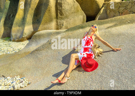 Attraente stile di vita bionda donna con un simpatico cane a prendere il sole su un masso di granito ad Anse Source d'Argent una delle più belle spiagge di La Foto Stock