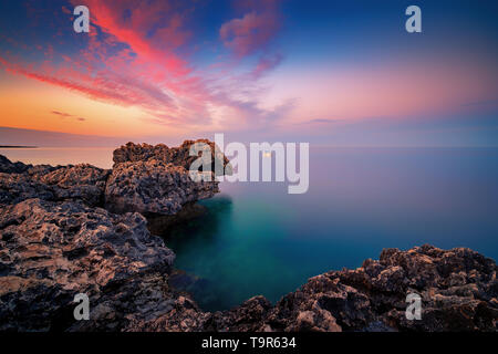 Un tramonto meraviglioso immagine di Cape Greco scogliere e rocce su un tramonto nella città di Paralimni, Cipro. Colorato di rosso, rosa e giallo skies con mare blu turchese. Foto Stock