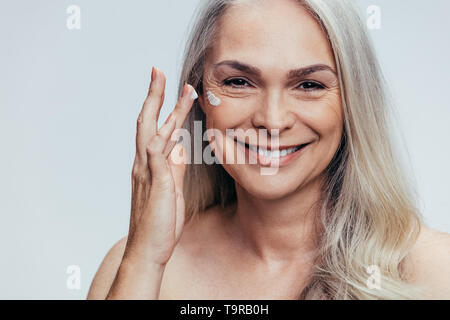 Sorridente metà adulto donna caucasica applicando anti invecchiamento la crema sul suo viso. Senior donna femmina di applicare una crema idratante sulla sua faccia contro uno sfondo grigio. Foto Stock