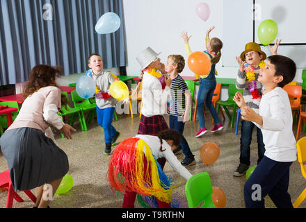Felice di ridere gli alunni della scuola primaria per divertirsi durante il festoso evento con il loro insegnante femminile, divertente con palloncini colorati Foto Stock