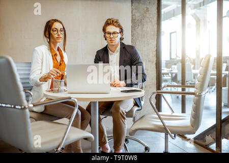 Giovane uomo e donna avente una conversazione aziendale durante la conferenza di piccole dimensioni, seduti a tavola rotonda nella sala riunioni