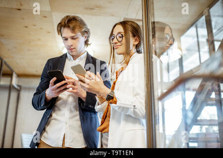 Uomo di affari e donna avente una conversazione mentre in piedi insieme con i telefoni intelligenti nell'ufficio moderno Foto Stock