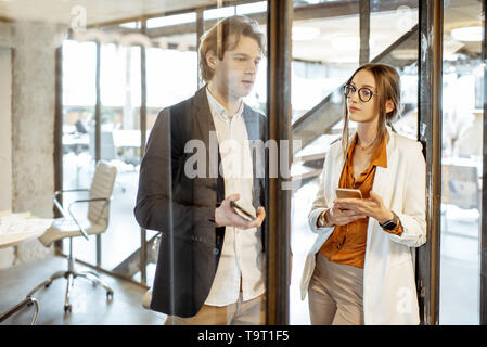 Uomo di affari e donna avente una conversazione mentre in piedi insieme con i telefoni intelligenti nell'ufficio moderno Foto Stock