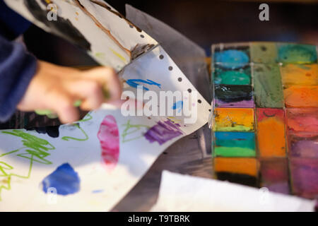 Le mani di un bambino rabberciato e taglio di un foglio di carta bianco con la pittura ad acquerello con un paio di forbici Foto Stock