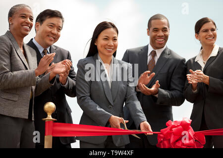 Sorridente di mezza età imprenditrice esegue la cerimonia del taglio del nastro di un nuovo sviluppo come i suoi colleghi guardano su felicemente e applaudire. Foto Stock