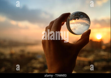 La mano trattiene Palla di vetro che riflette il Cielo di tramonto sulla città. Foto Stock