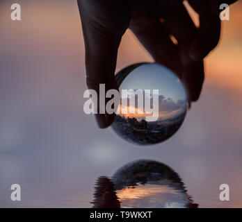 Mano trattiene Palla di vetro che riflette il Cielo di tramonto sulla città. Foto Stock