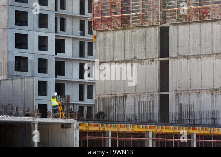 Il builder in tuta gialla fa un rilievo topografico contro lo sfondo di un muro in calcestruzzo, un multi-appartamento edificio in costruzione. Foto Stock