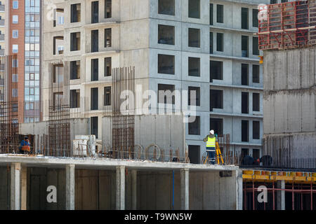 Il builder in tuta gialla fa un rilievo topografico contro lo sfondo di un muro in calcestruzzo, un multi-appartamento edificio in costruzione. Foto Stock