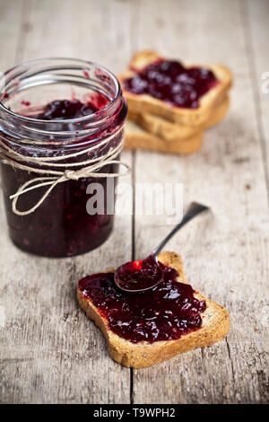 Cereali tostati le fette di pane e jar con fatti in casa frutti di bosco Confettura e closeup cucchiaio su tavola in legno rustico sfondo. Sweet Food per la prima colazione con Foto Stock