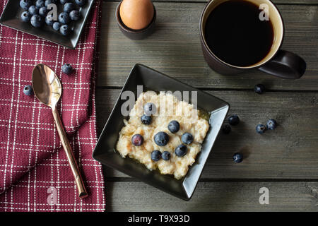 La colazione tazza di fiocchi d'avena, miele, burro e mirtilli con caffè e uova sode su un tavolo rustico con un cucchiaio e canovaccio Foto Stock