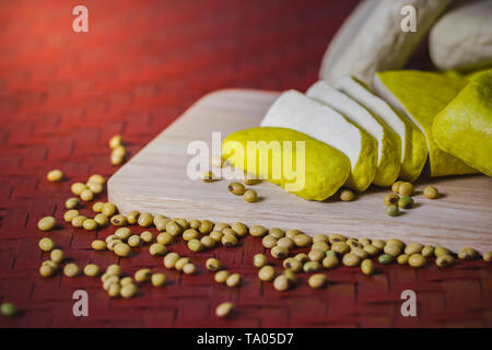 Il tofu è tagliato su un tagliere di legno e semi di soia sparsi sul tavolo. Cibo sano concetto. Foto Stock