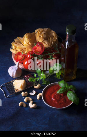 La tabella con gli ingredienti per fare il pesto di pomodoro. Pomodori, aglio, origano fresco e basilico erbe, bottiglia di olio d'oliva, alcune noci di acagiù, parmigiano, c Foto Stock