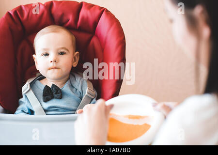 Alimentazione madre bambino seduto nella sedia in cucina. Guardando alla fotocamera. La maternità. La maternità. Foto Stock