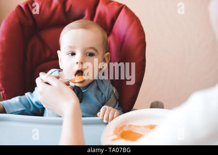 Baby boy 1 anno vecchio mangiare purea di mela seduto nella sedia in cucina. La madre del bambino di alimentazione. Infanzia. Foto Stock