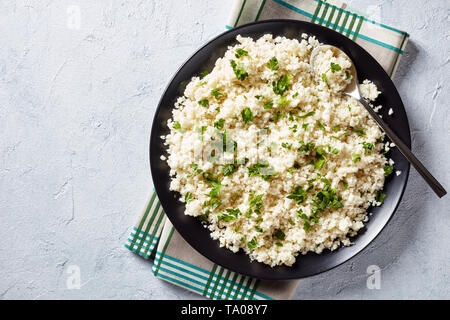 Il cavolfiore riso o cous cous mescolato con del prezzemolo tritato finemente in una ciotola nero su bianco in cemento Tabella, mangiare sano, basse calorie cibo, horizonta Foto Stock