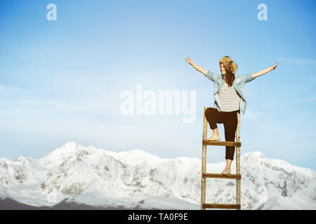 Ragazza Felice sorge su scala in legno con braccia sollevate contro le montagne nevose Foto Stock