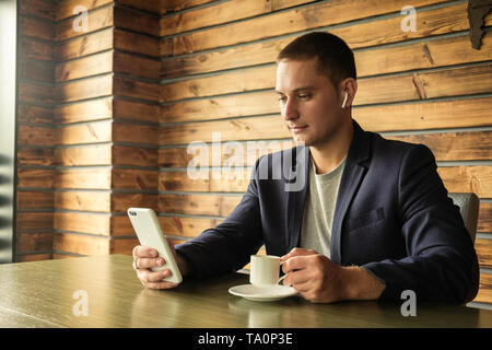 Imprenditore in ascolto su di auricolari per il suo telefono cellulare mentre bevendo una tazza di caffè al suo banco in una vista ravvicinata Foto Stock