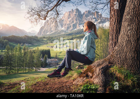 Bella giovane donna in occhiali da sole e giacca blu è seduto sulla collina sotto l'albero al tramonto. Molla colorato paesaggio con ragazza sportivo, meado Foto Stock