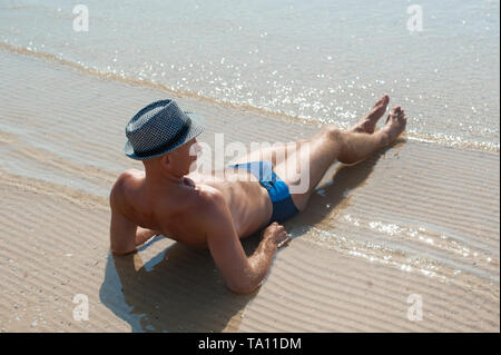Elegante giovane maschio modello uomo disteso sulla spiaggia di sabbia di indossare hipster estate hat godendo di viaggio estivo vacanza vicino al mare Foto Stock