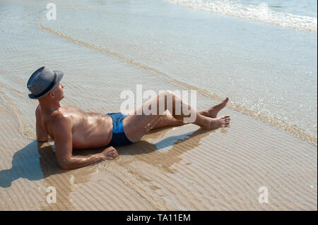 Elegante giovane maschio modello uomo disteso sulla spiaggia di sabbia di indossare hipster estate hat godendo di viaggio estivo vacanza vicino al mare Foto Stock