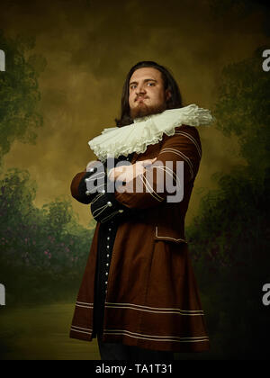 Giovane uomo come un cavaliere medievale su studio scuro dello sfondo. Ritratto in chiave di basso del modello maschile in costume retrò. Guardando gravi. Le emozioni umane, compari Foto Stock