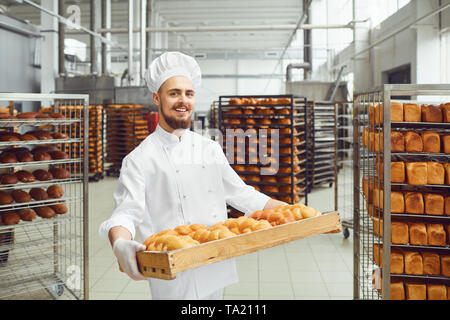 Sorridente baker in bianco uniforme con vassoi di pane fresco di costi nella produzione di panetteria. Foto Stock